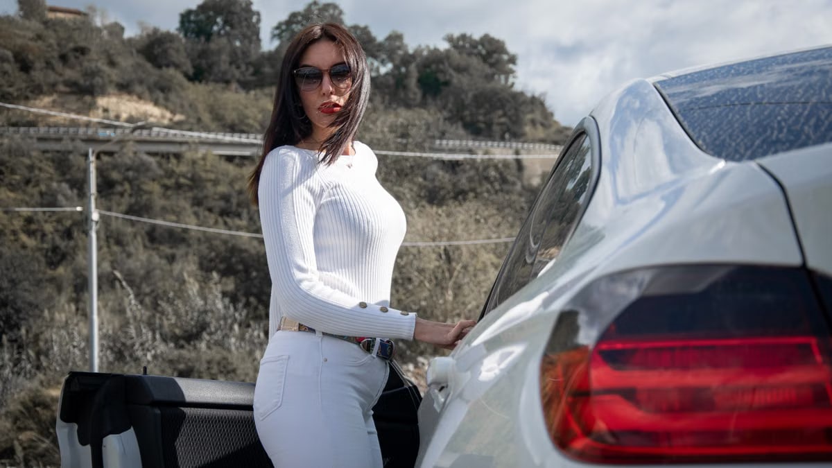 Woman in sunglasses posing beside a car on a sunny day, showcasing elegance and fashion.